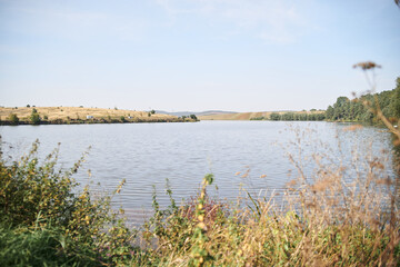 Amazing lake landscape with dry grass and green trees in autumn.Light blue sky over pond. Nature protection concept. Rural scene in countryside. Fish farming basin in the village.