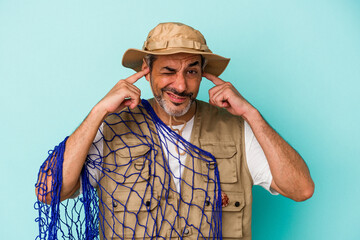 Middle age caucasian fisherman holding net isolated on blue background  covering ears with hands.