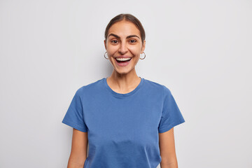 Studio shot of happy dark haired young woman smiles broadly expresses positive emotions dressed in casual blue t shirt isolated over white background. Cheerful female model reacts on funny news