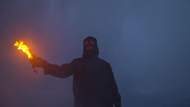 A man in a hood and balaclava holds a burning molotov cocktail in his hand