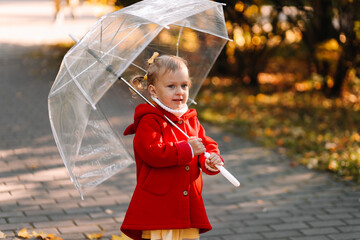 A cheerful little girl child in a warm red coat holds a transparent umbrella, has fun with fall...