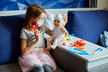 A little girl in medical mask sits on the sofa, plays a doctor, treats to the doll