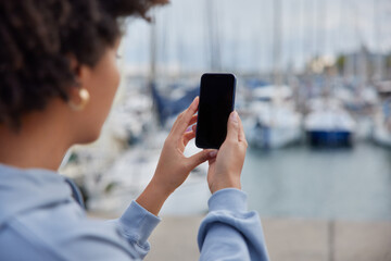 Unrecognizable woman takes photo of harbor poses on pier holds modern mobile phone with blank...