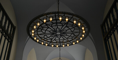 Round ornamental chandelier in black with light bulbs hanging from ceiling behind open gates.