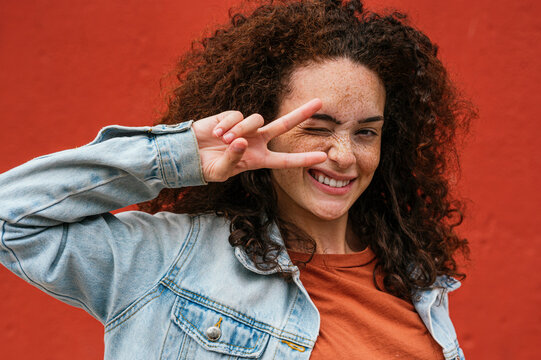 Playful young woman showing peace sign while winking in front of red wall