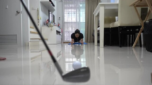 Asian man hitting a golf ball past an Asian woman doing planking exercises at home at leisure while golf courses are closed due to COVID-19.