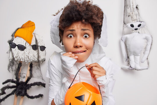 Scared worried curly haired African American woman holds carved pumpkin afraids of spooky creatures around bites lips dresses up in ghost cotume isolated over white background. Halloween concept