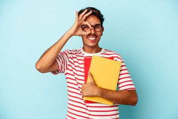 Young mixed race student man holding books isolated on blue background excited keeping ok gesture on eye.