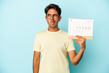 Young mixed race man holding calendar isolated on blue background confused, feels doubtful and...
