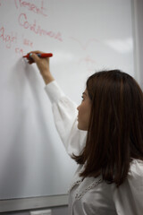 Profile view of a cute young brunette english teacher in white blouse writing on a board and giving a class.