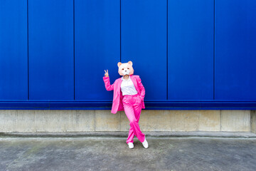 Woman wearing vibrant pink suit and bear mask making peace sign in front of blue wall