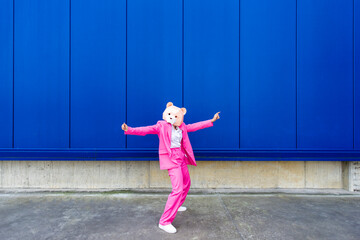Woman wearing vibrant pink suit and bear mask standing with outstretched arms in front of blue wall