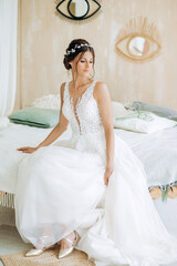Fototapeta na wymiar Cute bride in a white dress sitting on the bed in a stylish room.