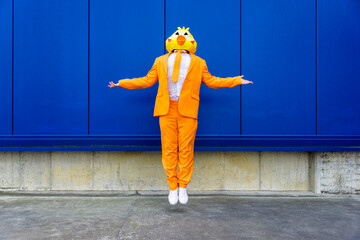 Man wearing vibrant orange suit and bird mask levitating in front of blue wall