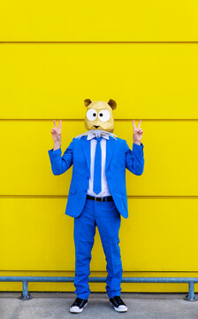 Man wearing vibrant blue suit and rodent mask making peace sign gestures in front of yellow wall