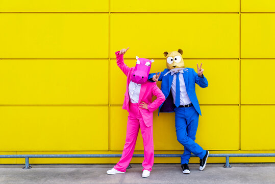 Man and woman wearing vibrant suits and animal masks posing together in front of yellow wall