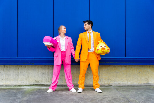 Man and woman wearing vibrant suits standing together in front of blue wall with animal masks in hands