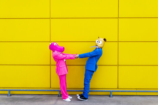 Man and woman wearing vibrant suits and animal masks holding hands in front of yellow wall