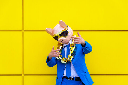 Man wearing vibrant blue suit, pig mask and large golden chain posing against yellow wall