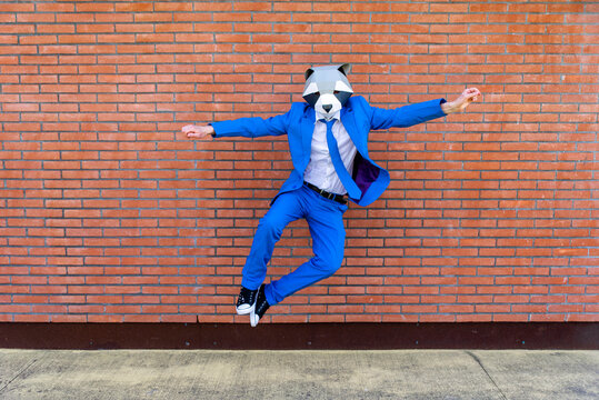 Man wearing vibrant blue suit and raccoon mask jumping against brick wall