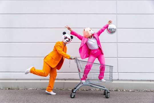 Man and woman wearing vibrant suits and animal masks messing around with shopping cart and disco ball