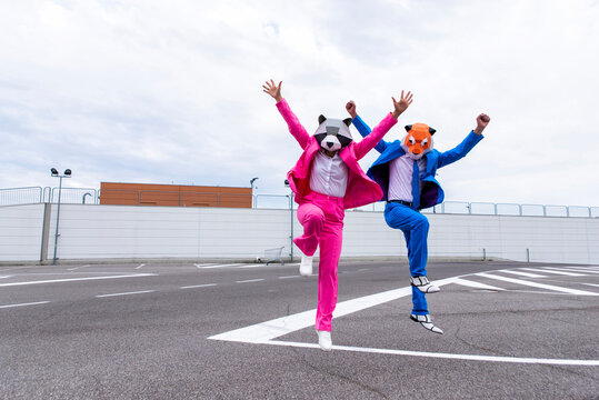 Man And Woman Wearing Vibrant Suits And Animal Masks Jumping Side By Side In Empty Parking Lot