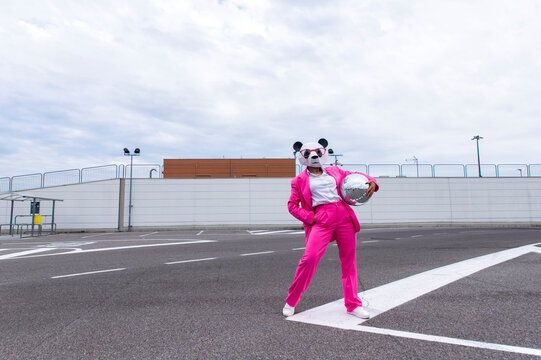 Woman wearing vibrant pink suit and panda mask posing in empty parking lot with disco ball in hand