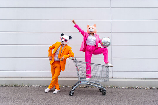 Man and woman wearing vibrant suits and animal masks&nbsp;posing with shopping cart and disco ball in front of white wall