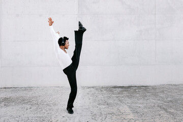 Flexible male dancer stretching leg while practicing urban dance against white wall