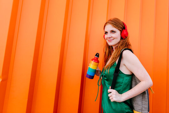 Woman with headphones and backpack holding multi colored bottle by orange wall