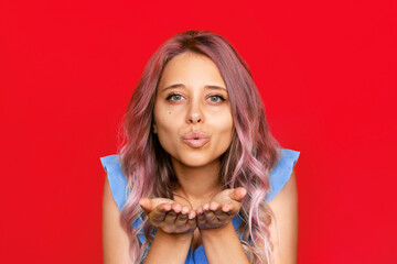 A young caucasian beautiful blonde woman with wavy pink hair sending air kiss with open palms looking at the camera isolated on a bright color red background. Pretty charming girl blows kiss