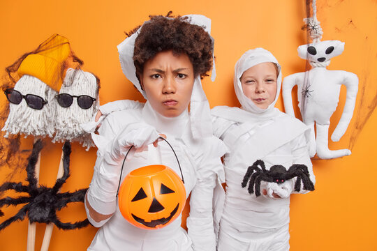 Displeased Adult Woman And Child Wear Scary Costumes For Halloween Party Pose With Carved Pumpkin And Spooky Spider Decorates House With Monsters And Different Horrible Creatures. Fearful Holiday