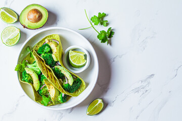 Green vegan tacos with broccoli, avocado and green flat bread in white bowl, white marble...