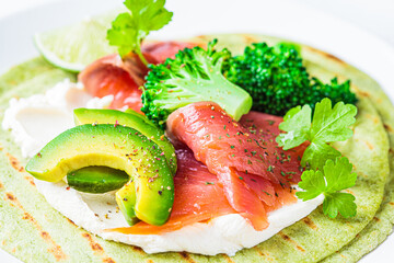 Green tortilla with broccoli, avocado, cream cheese and salted salmon on white marble background. Vegetarian fast food.
