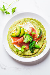 Green tortilla with broccoli, avocado, cream cheese and salted salmon on white marble background. Vegetarian fast food.