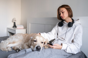 An upset girl sits with a dog on the bed, looks at the phone and listens to sad music. Teenage depression.