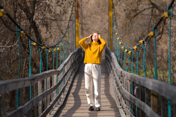 A red-haired girl stands on a wooden bridge in an orange sweater and looks at the autumn sun. Relaxed young woman walks in the forest.