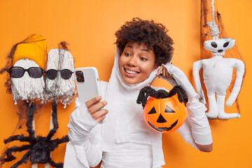 Glad young ethnic woman with curly hair makes video call via smartphone shows carved pumpkin with...