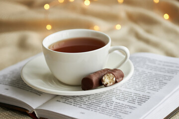 White cup of tea with chocolate sweets on a n open book and beige plaid in the Christmas lights.