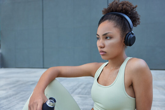 Horizontal shot of thoughtful young Afro American woman has sweat after intensive cardio training takes break drinks refreshing water from bottle dressed in sportswear listens motivational music