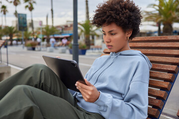 Serious Afro American woman tourist sits on bench in city concentrated at screen of digital tablet...