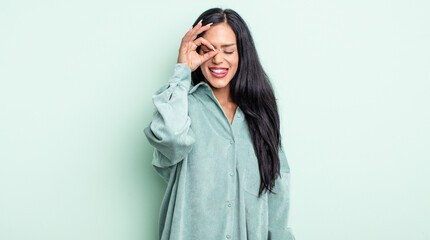 pretty hispanic woman smiling happily with funny face, joking and looking through peephole, spying...