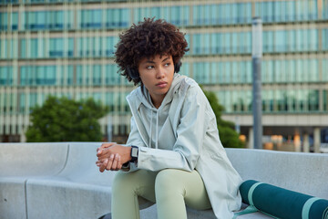 Fototapeta na wymiar Thoughtful young Afro American woman rests after hard workout looks away pensively keeps hands together sits tired poses against urban background with fitness mat listens music in headphones