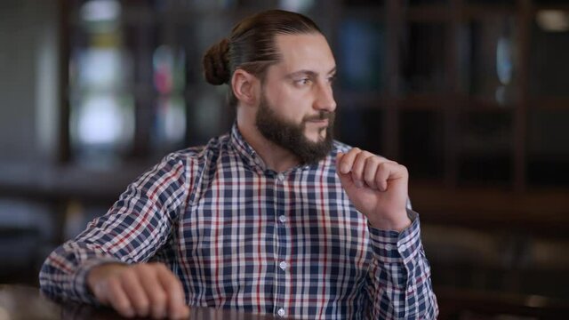 Thoughtful handsome Caucasian man sitting at bar counter waiting looking around. Portrait of bearded confident boyfriend waiting for a date in pub indoors. Punctuality and relationship