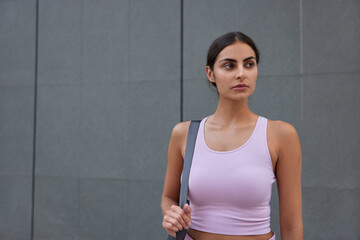 Fototapeta na wymiar Horizontal shot of thoughtful sportive woman dressed in active wear carries karemat has fitness mat going to have workout poses against grey wall with blank space for your promotional content