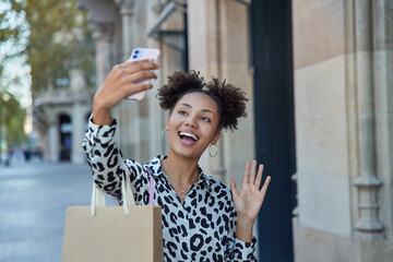 Shopping sale and lifestyle concept. Positive Afro American teenage girl holds smartphone waves hello makes online call communicates with friend carries paper bag returns from mall has good time