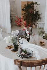 Elegant aesthetic Christmas dinner serving concept. Bright living room interior with chair, table with beige linen table cloth, berries bouquet. Christmas celebration evening