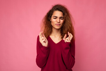 Photo of young emotional attractive brunette curly woman with sincere emotions wearing trendy pink sweater isolated over pink background with empty space and holding fingers crossed for good luck