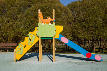 A wooden slide of bright blue and yellow color against a background of green trees on a clear sunny day. Playgrounds, sports, health entertainment.