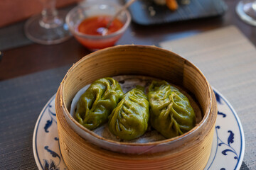Oriental green dumplings in a traditional bamboo steamer, background chili sauce at restaurant.